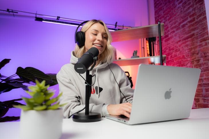 Mujer sentada detrás de escritorio blanco sonriendo con un micrófono y una computadora Apple al frente